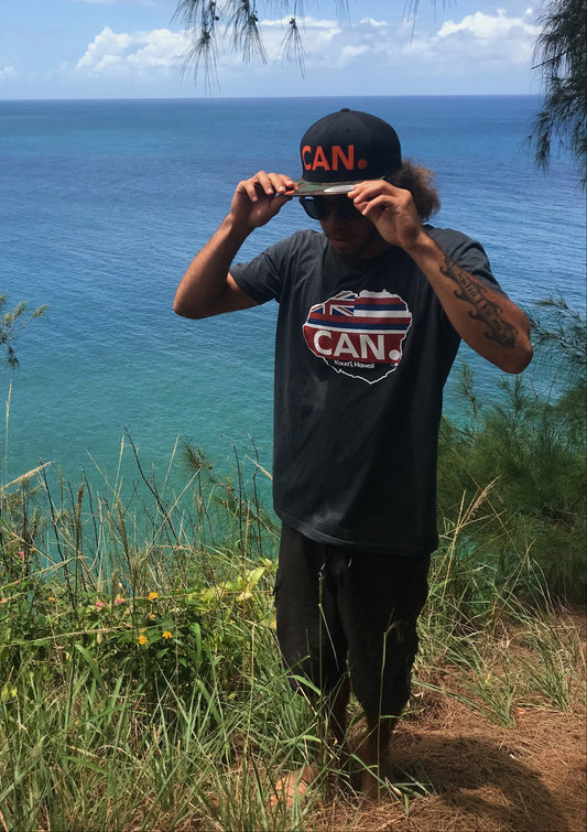 Person wearing a cap and t-shirt with a logo, standing on a grassy hill overlooking the ocean.